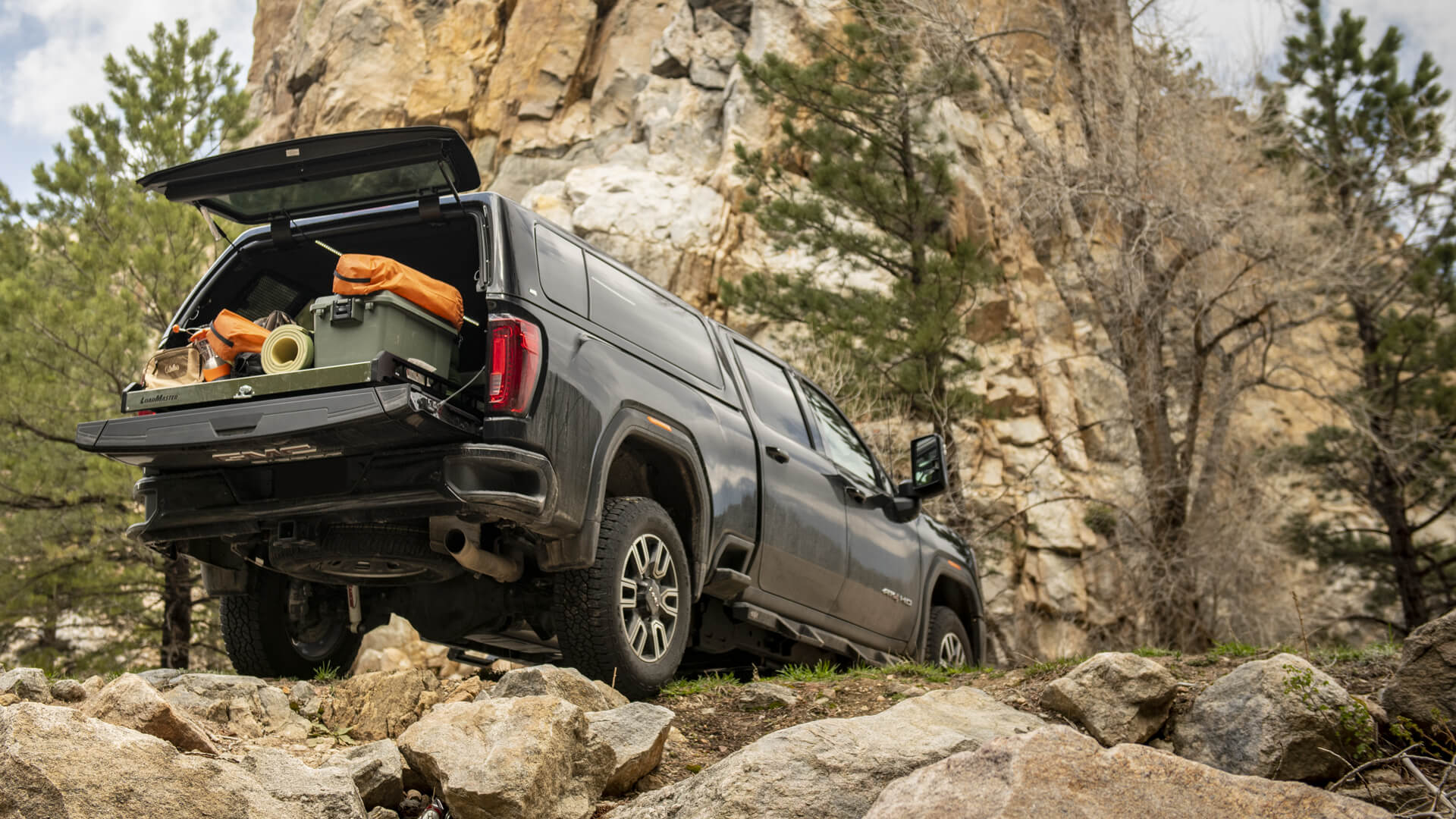 Black GMC Sierra with an ATC truck cap containing camping supplies and perched on a rocky road in a forest under an overcast sky
