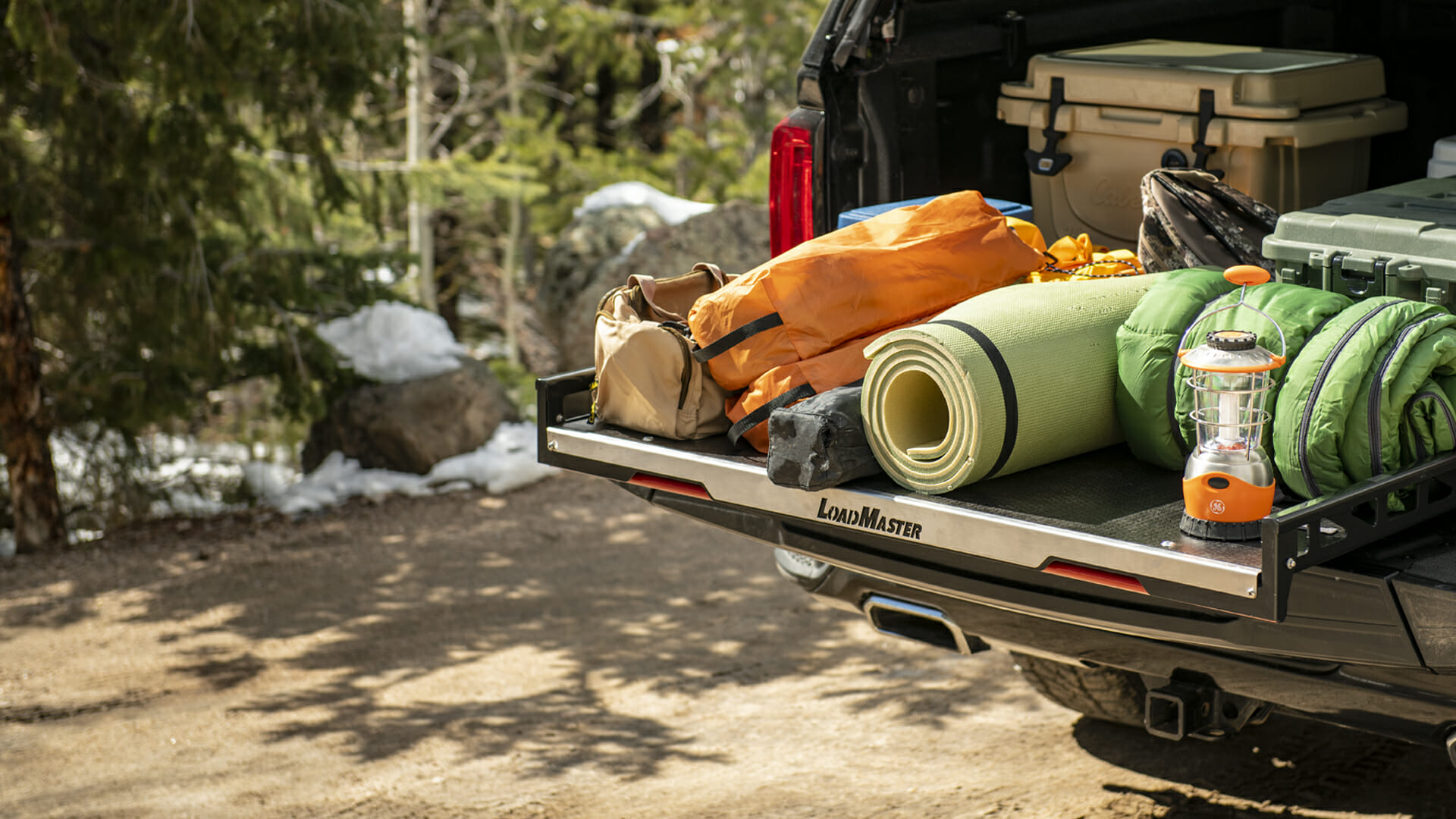 Loadmaster Utility Tray in a black pick up truck containing a lantern, sleeping bag, mat, boxes, foldable chairs, a backpack, and other camping supplies on a dirt road in a snowy forest